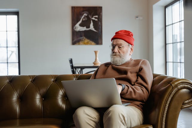 Elderly man wearing red beanie using laptop on sofa at home.