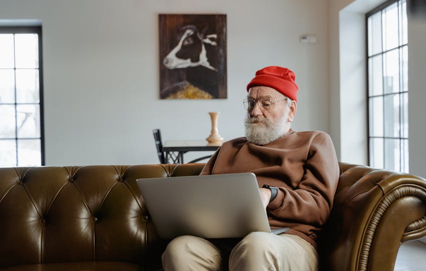 Elderly man wearing red beanie using laptop on sofa at home.