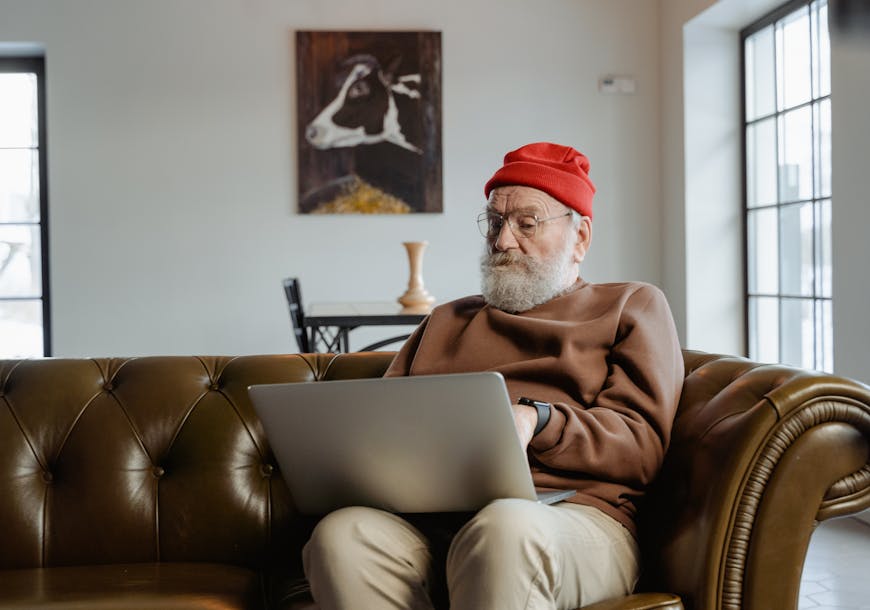 Elderly man wearing red beanie using laptop on sofa at home.