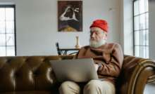 Elderly man wearing red beanie using laptop on sofa at home.